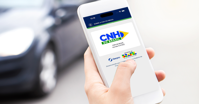 Hand holding a smartphone displaying the official CNH do Brasil app logo, with a car blurred in the background